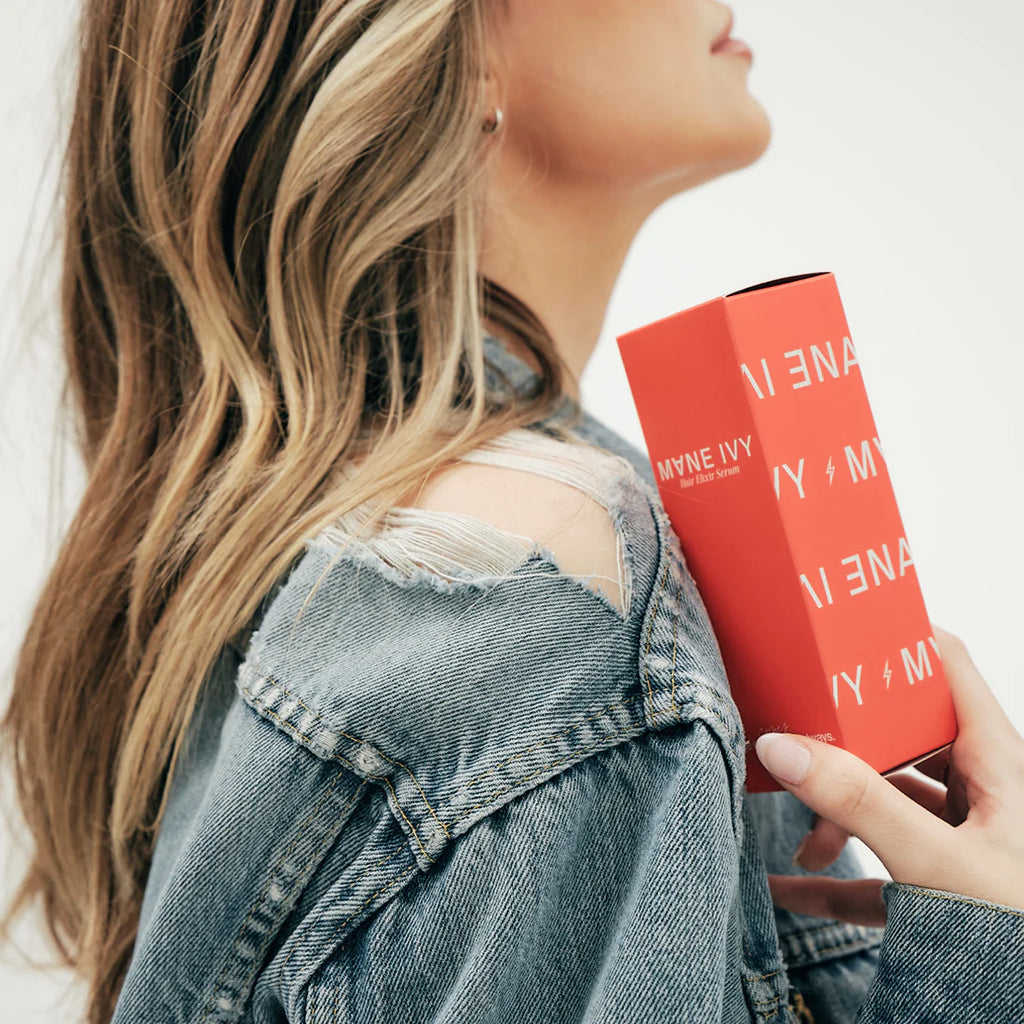 This shows a blonde woman holding the hair elixir serum box. The box is bright red and has the Mane Ivy logo on it. The serum is meant to moisturize your hair and help with breakage and heat protection. 